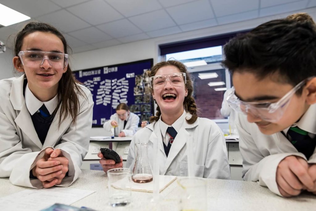 A group of pupils of secondary school age wearing lab coats and laughing in a science classroom