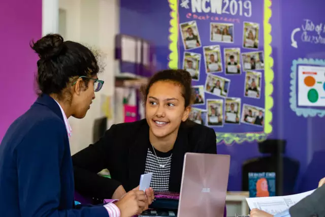 Two teenage girls talking in a classroom setting