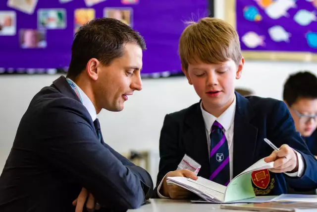 A teacher knelt down next to a pupil at their desk and to help with their secondary school work