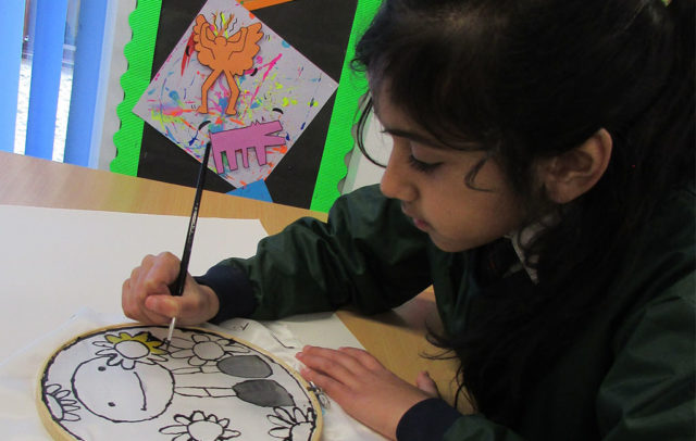 School girl sat at a desk, holding a paintbrush and painting a picture